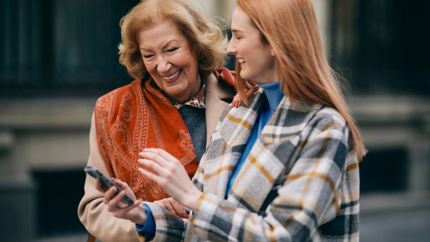 An older woman and a young woman laughing together over a phone on a city sidewalk